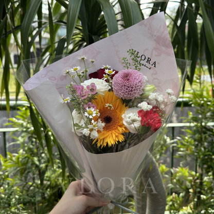 Bouquet of flowers wrapped in white paper with 'VLORA' branding, held against a green foliage background.
